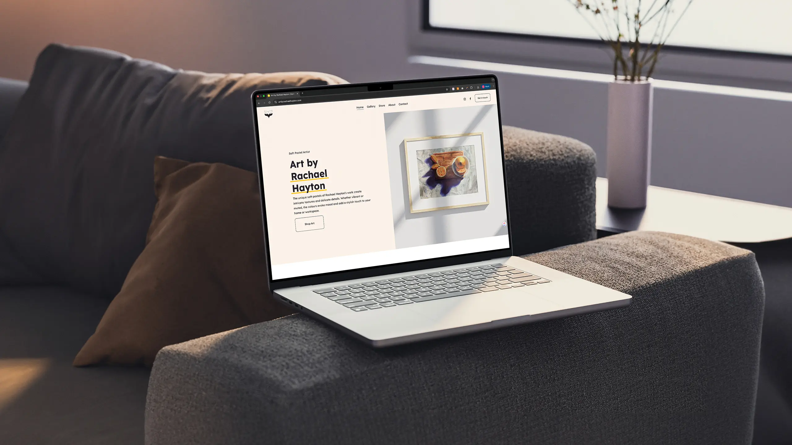 An Apple MacBook placed on the arm of a grey couch displaying a website with a cream coloured background, black text and a photo of a framed artwork